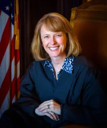 Female judge smiling, seated with American flag background.